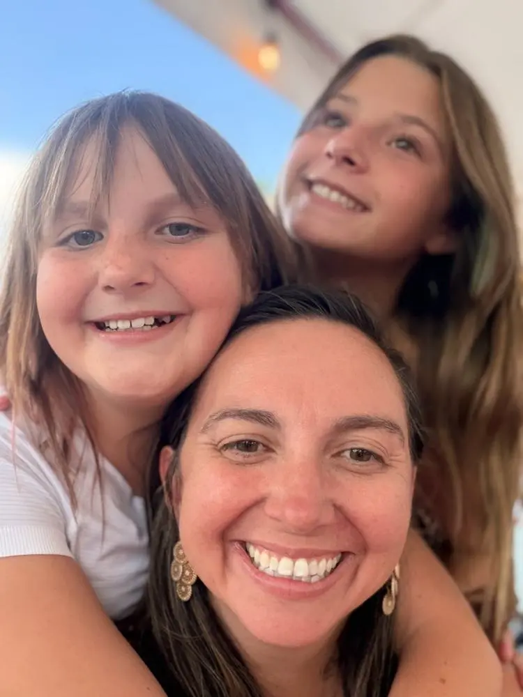 Val smiling with her two daughters hugging her from behind